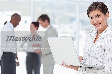 Smiling young saleswoman with her laptop and colleagues behind her