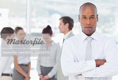 Confident young salesman with arms crossed and colleagues behind him