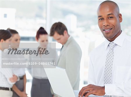 Smiling young salesman with his laptop and associates behind him