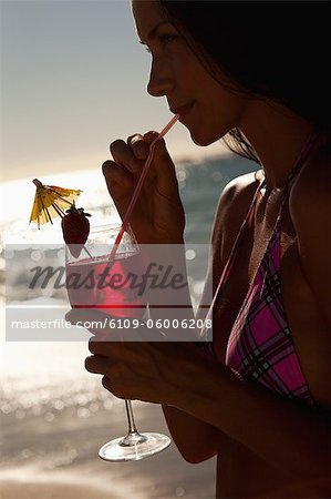 Woman wearing a bikini while drinking a cocktail as she stands on a beach with the sun shining on one side of her body