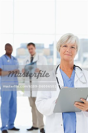 Mature doctor with clipboard and two talking colleagues behind her