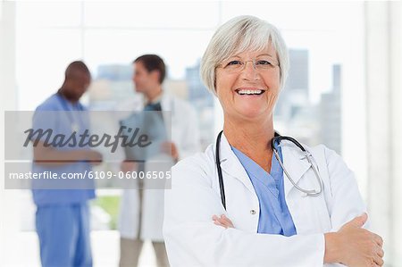 Smiling mature doctor with folded arms and two colleagues behind her