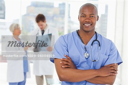 Smiling doctor in scrubs with arms folded and two colleagues behind him