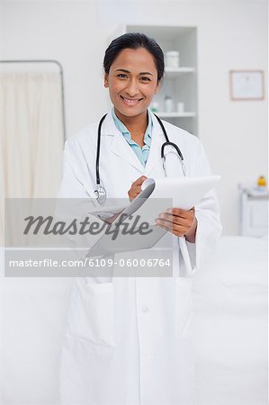 Young doctor writing on a clipboard while smiling and standing in a hospital room