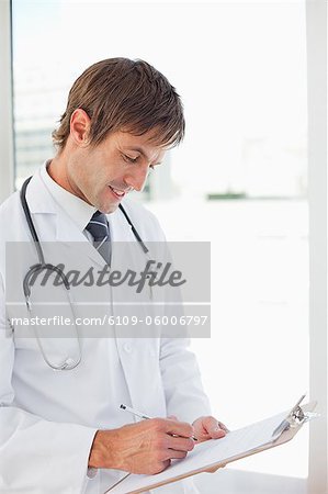 Smiling doctor working with his clipboard while standing in front of a window