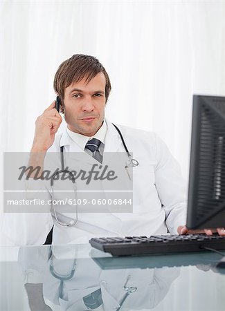 Thoughtful doctor using his mobile phone while raising an eyebrow and sitting at his desk