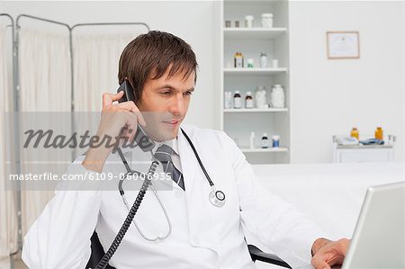 Serious doctor working on his laptop while using his phone