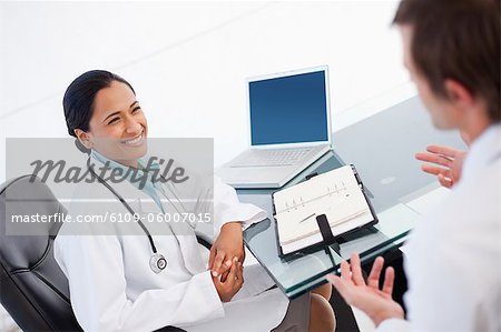 Happy doctor listening to a co-worker while sitting at a desk with a diary