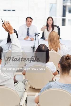 Man raising his hand as he sits in an audience while watching two business people who are sitting behind a table