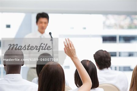 Woman raises her hand while sitting with her colleagues while watching a businessman