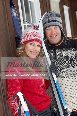 Couple standing by log cabin, holding skis.