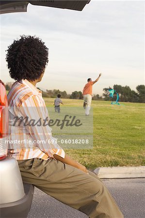 Mother sitting on open car boot watching father and son fly kite