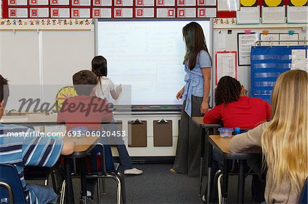 Female Teacher standing  by whiteboard and assisting schoolgirl