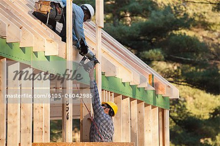 Carpenter handing nail gun to another carpenter on roof