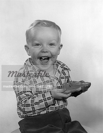 1950s LAUGHING BOY WEARING A PLAID SHIRT WITH JAM ON HIS FACE & MOUTH HOLDING A SLICE OF BREAD WITH BOTH HANDS LOOKING AT CAMERA