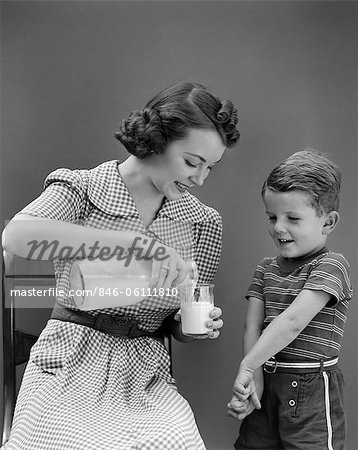 1940s WOMAN MOTHER SITTING POURING GLASS OF MILK FOR BOY SON STANDING BESIDE HER