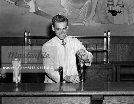 ANNÉES 1940 SOURIANT SODA JERK HABILLÉ EN CHEMISE CRAVATE & WHITE BLOUSE DEBOUT DERRIÈRE LE COMPTOIR DE FONTAINE REMPLISSAGE VERRE EN REGARDANT LA CAMÉRA