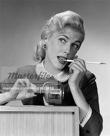 1950s DISTRACTED BLOND WOMAN OFFICE WORKER AT PENCIL SHARPENER LOOKING OFF TO SIDE WITH PENCIL IN MOUTH