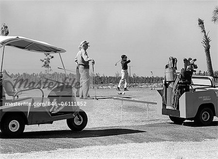 1960s THREE MEN PLAYING GOLF TEEING OFF FROM TEE WITH DRIVER NEAR GOLF CARTS OUTDOOR