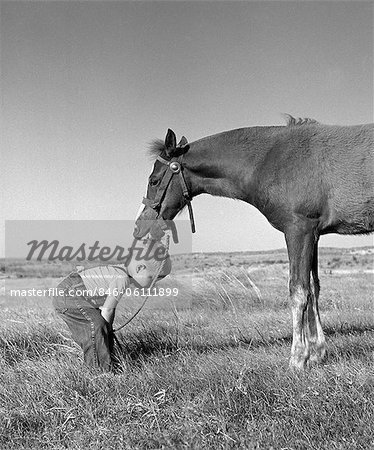 1950s SIDE VIEW OF LITTLE BLOND BOY IN FIELD WEARING JEANS & SUSPENDERS BENDING DOWN WITH HORSE LICKING TOP OF HEAD