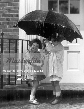 1920s PAIR OF LITTLE GIRLS STANDING ON FRONT STOOP UNDER UMBRELLA IN POURING RAIN