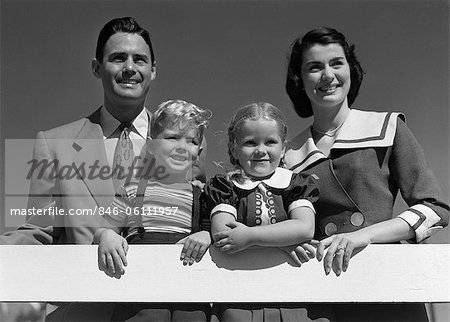 1950s PORTRAIT SMILING FAMILY FATHER MOTHER DAUGHTER SON STANDING TOGETHER BEHIND WHITE FENCE OUTDOOR