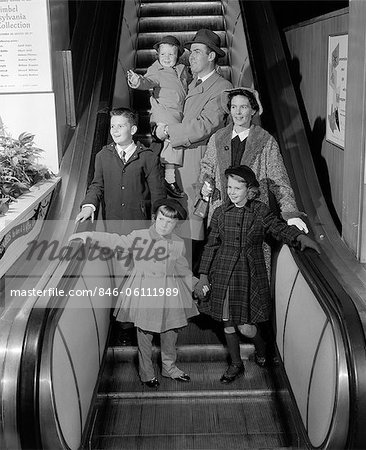 1950s SMILING FAMILY OF 6 IN WINTER COATS GOING DOWN STORE ESCALATOR