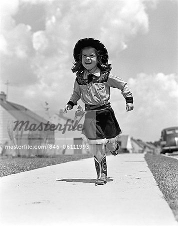 ANNÉES 1950 GIRL IN COWGIRL COSTUME EN COURS D'EXÉCUTION SUR LE TROTTOIR VERS LA CAMÉRA