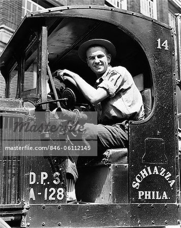 1940s TRUCK DRIVER LOOKING AT CAMERA IN WORK SHIRT & HAT POSED BEHIND WHEEL OF TRUCK WITH OPEN SIDE NO DOOR