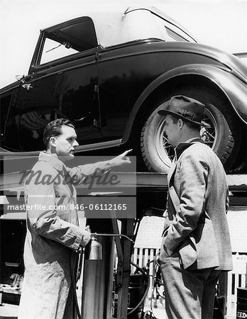 1930s SERVICE STATION ATTENDANT IN COVERALLS HOLDING OIL CAN POINTING AT PROBLEM CONVERTIBLE ON LIFT TALKING TO OWNER
