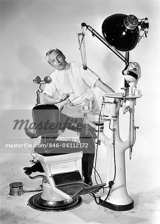 1950s MAN DENTIST HOLDING GLASSES SPEAKING FOOT PROPPED UP ON DENTAL CHAIR AND EQUIPMENT LOOKING AT CAMERA