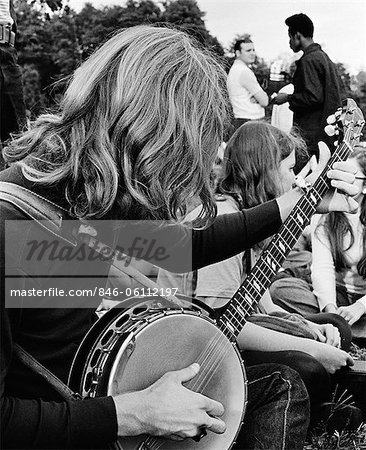 1970s SIDE VIEW OF HIPPIE TEEN OUTDOOR GATHERING WITH LONG-HAIRED BOY IN FOREGROUND PLAYING BANJO