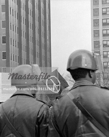 1970s BACK VIEW OF TWO POLICEMAN IN HELMETS AT DEMONSTRATION WITH PEACE SIGN IN BACKGROUND FRAMED BETWEEN THEIR HEADS