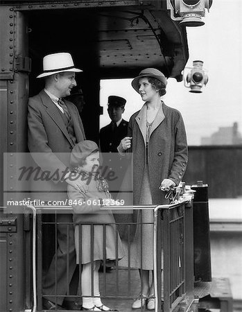 1930s FAMILY OF THREE STANDING AT RAILING AT BACK OF TRAIN CAR WITH PORTERS IN BACKGROUND