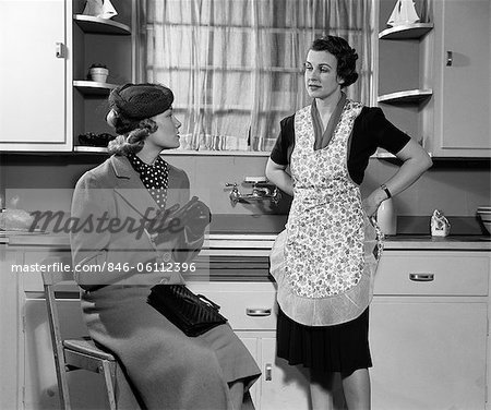 ANNÉES 1930 1940 DEUX FEMMES ASSISES EN CUISINE PARLER UNE FEMME AU FOYER AU FOYER EN TABLIER DANS UN MANTEAU ET CHAPEAU VISITER ENSEMBLE GOSSIP