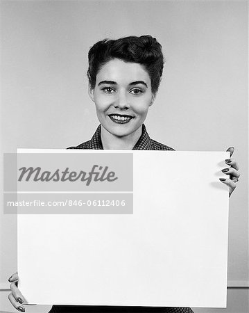 1950s WOMAN HOLDING BLANK SIGN IN FRONT OF CHEST SMILING INDOOR