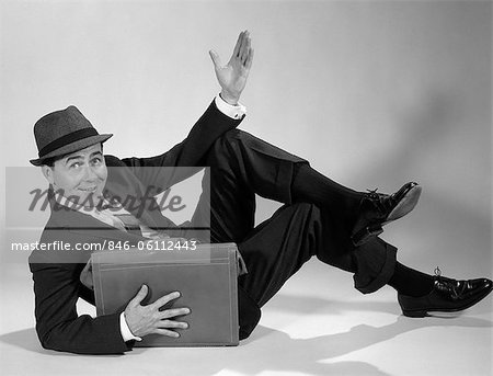 1960s BUSINESSMAN RECLINING PROPPED ON ELBOW HOLDING BRIEFCASE OTHER HAND UP IN THE AIR LOOKING AT CAMERA