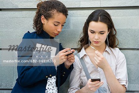 Young women together outdoors, text messaging and listening to MP3 player