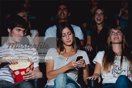 Woman using cell phone in movie theater, man looking over with annoyed expression