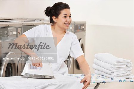 Happy young employee ironing clothes while looking away in Laundromat