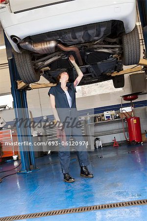 Young female mechanic working while standing under lifted car in workshop