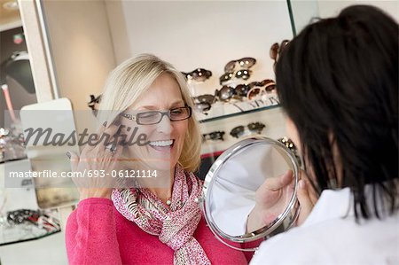 Cheerful senior woman trying on glasses while optician holding mirror