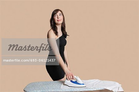 Beautiful young woman looking up while ironing shirt over colored background