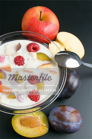 Glass bowl filled with yogurt mixed with fruit pieces arranged with spoon and some fruits around close-up  isolated on black background.