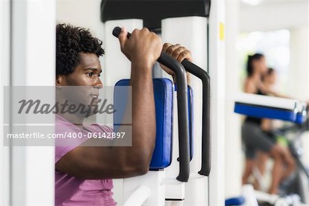 Young black man exercising pectoral muscles in fitness club, with people working out on cyclette in background