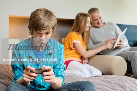 Teenage boy using mobile phone with his father and sister using digital tablet in background Teenage boy using mobile phone with his father and sister using digital tablet in background