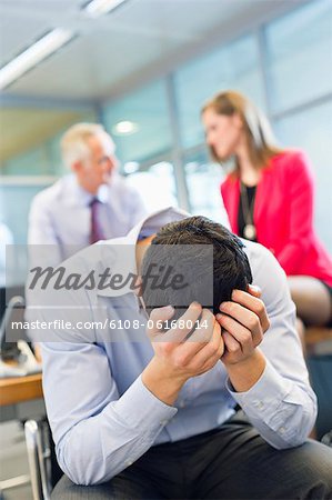 Male executive looking worried in an office with his colleagues discussing in the background