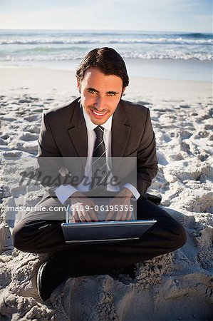 Smiling businessman sitting cross-legged in front of the sea with a laptop