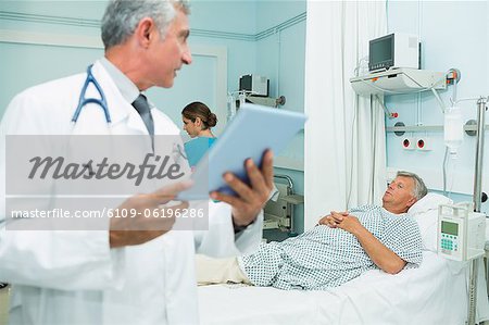 Male doctor using a tactile tablet while looking at a patient in a bed ward