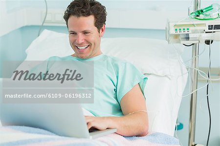 Happy male patient typing on a laptop while lying on a bed